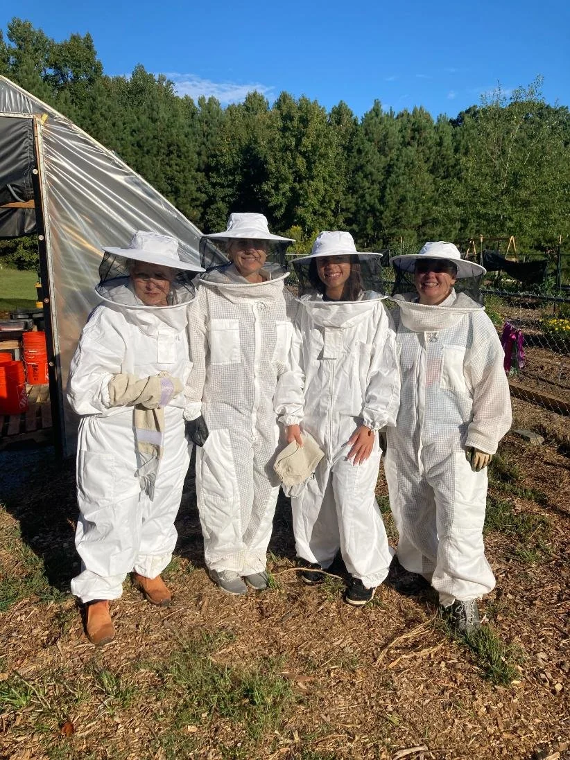 Four women wearing white beekeeping suits and hats with veils standing outdoors in front of a greenhouse and trees on a sunny day.