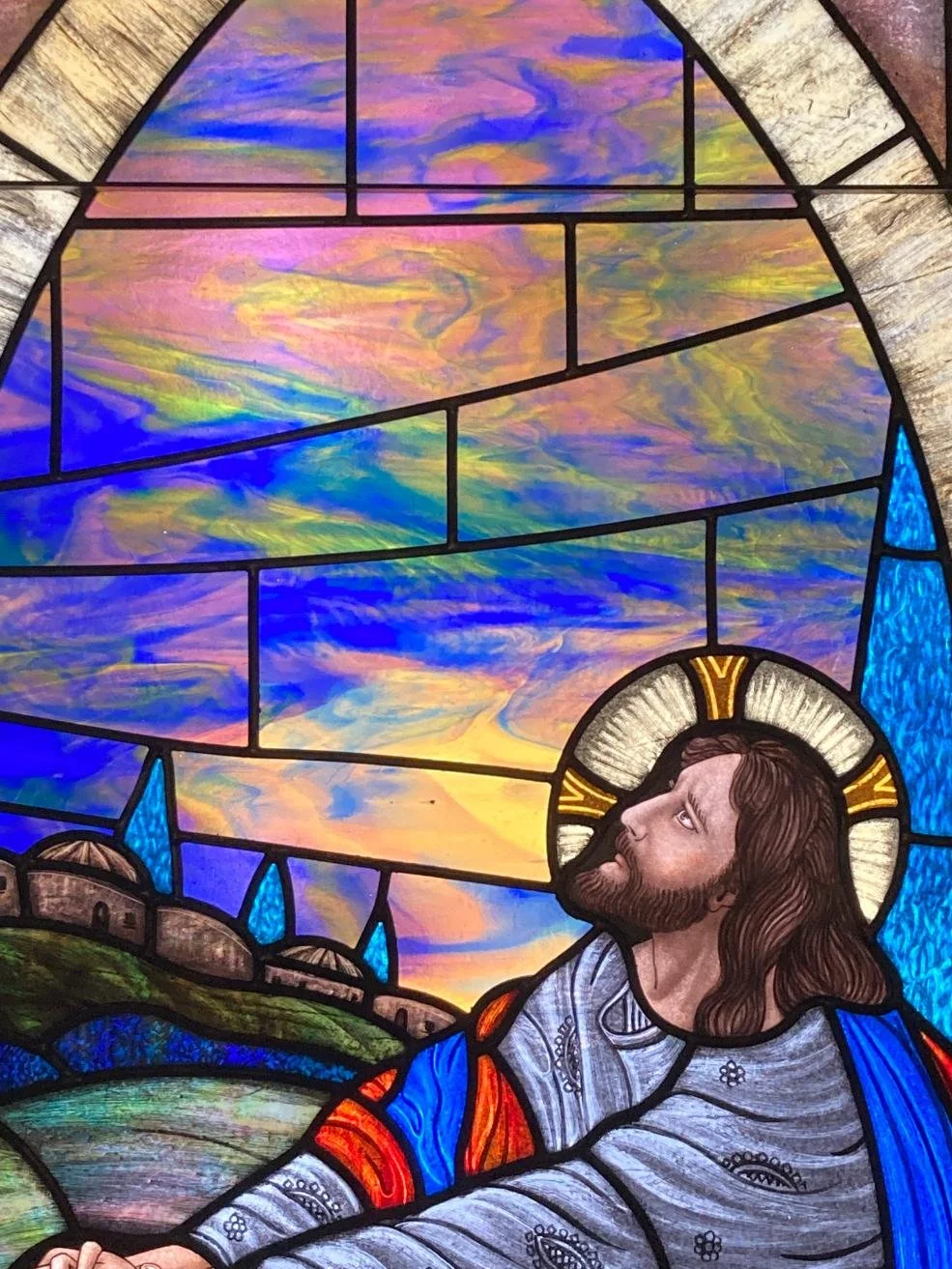 Stained glass window depicting Jesus Christ looking upward with a sunset sky and a town in the background.