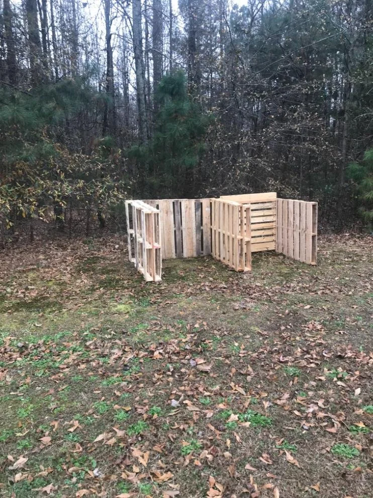 A small wooden fence constructed from repurposed pallets on a grassy area with fallen leaves, surrounded by trees and bushes in the background.