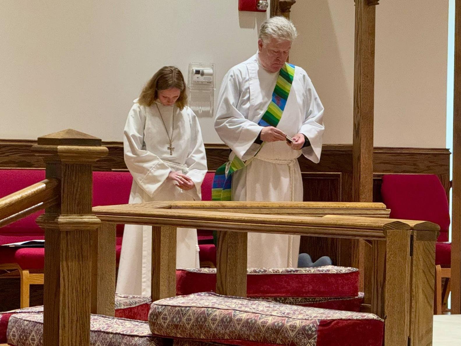 Two people dressed in white robes standing in a church, with their heads bowed and hands clasped or holding small objects, possibly during a prayer or religious ceremony.