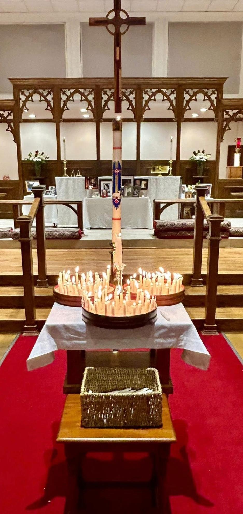 A candlelit memorial with lit candles arranged around a tall decorated candle, set in a church with wooden altar and cross, and framed photographs on the altar table.