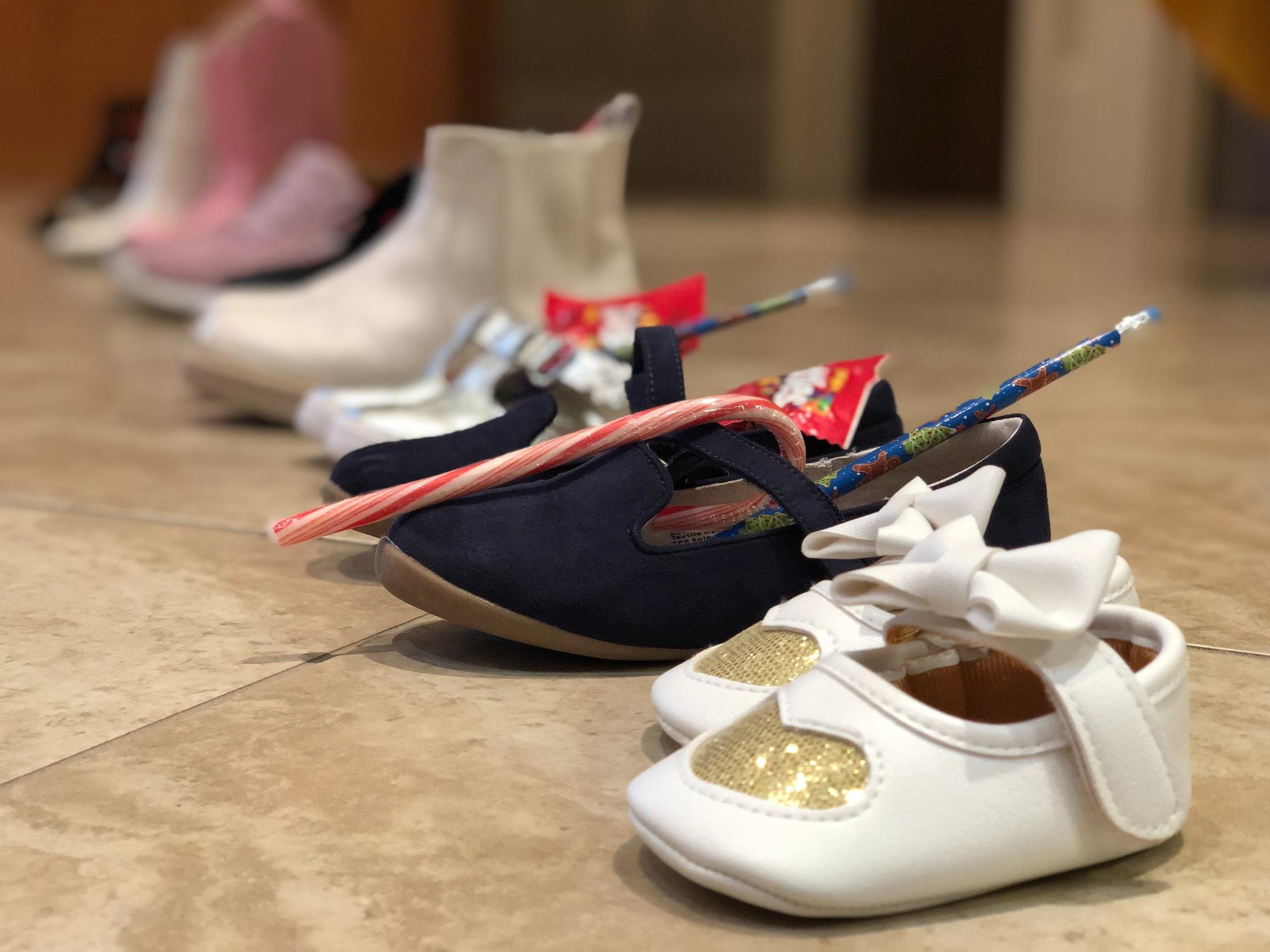 A row of small children's shoes and accessories lined up on a tiled floor, including white and navy shoes with bows, gold and white shoes, and various colorful shoes and socks generally used for infants.