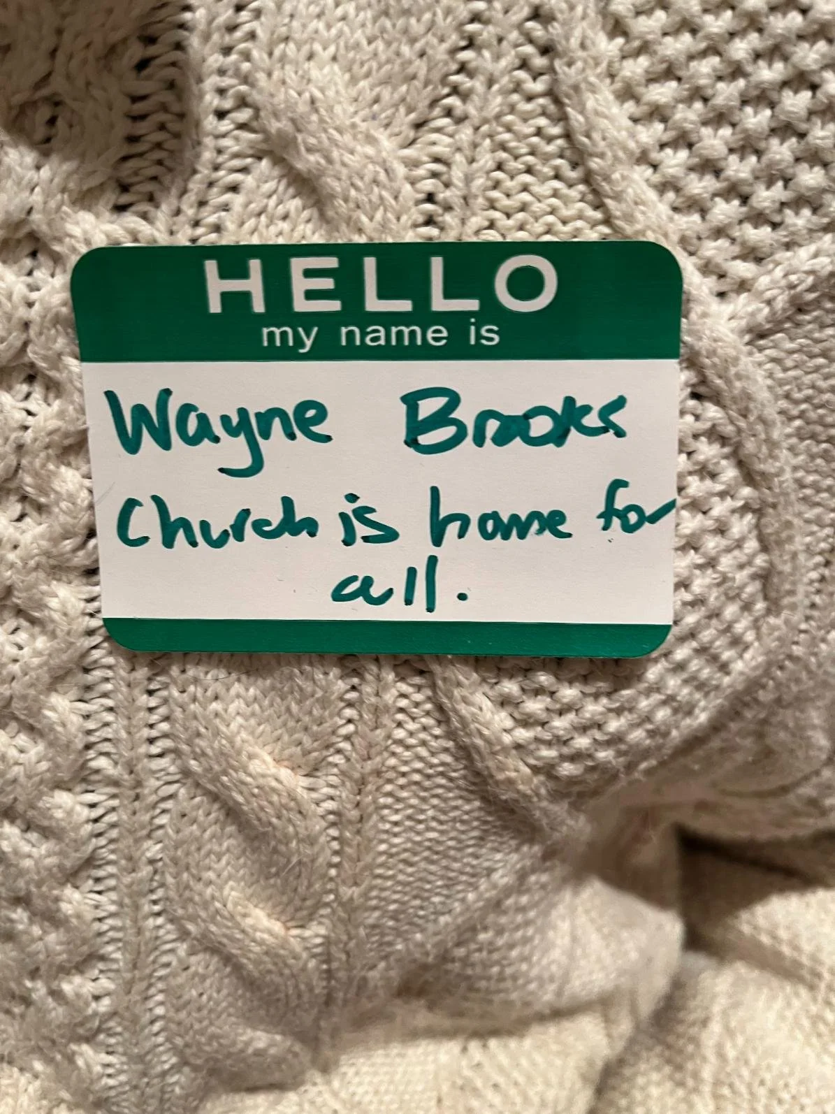 A name tag on a knitted white sweater reads, "Hello my name is Wayne Brooks. Church is home for all."