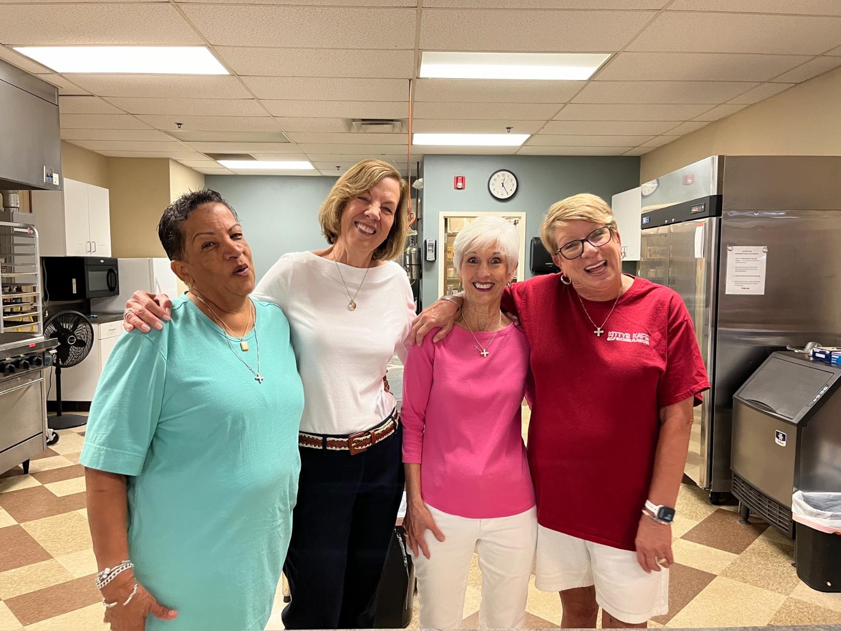 Four women standing together in a kitchen, smiling and posing for the photo, with one woman in a turquoise shirt, a second in a white shirt, the third in a pink shirt, and the last in a red shirt.