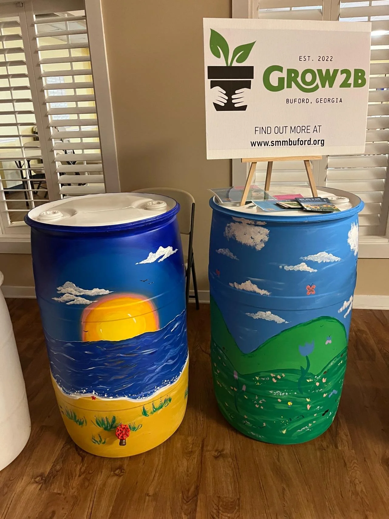Two painted plastic barrels, one depicting a sunset over the ocean and the other a green hillside with flowers, at a Grow2B organization event in Buford, Georgia.