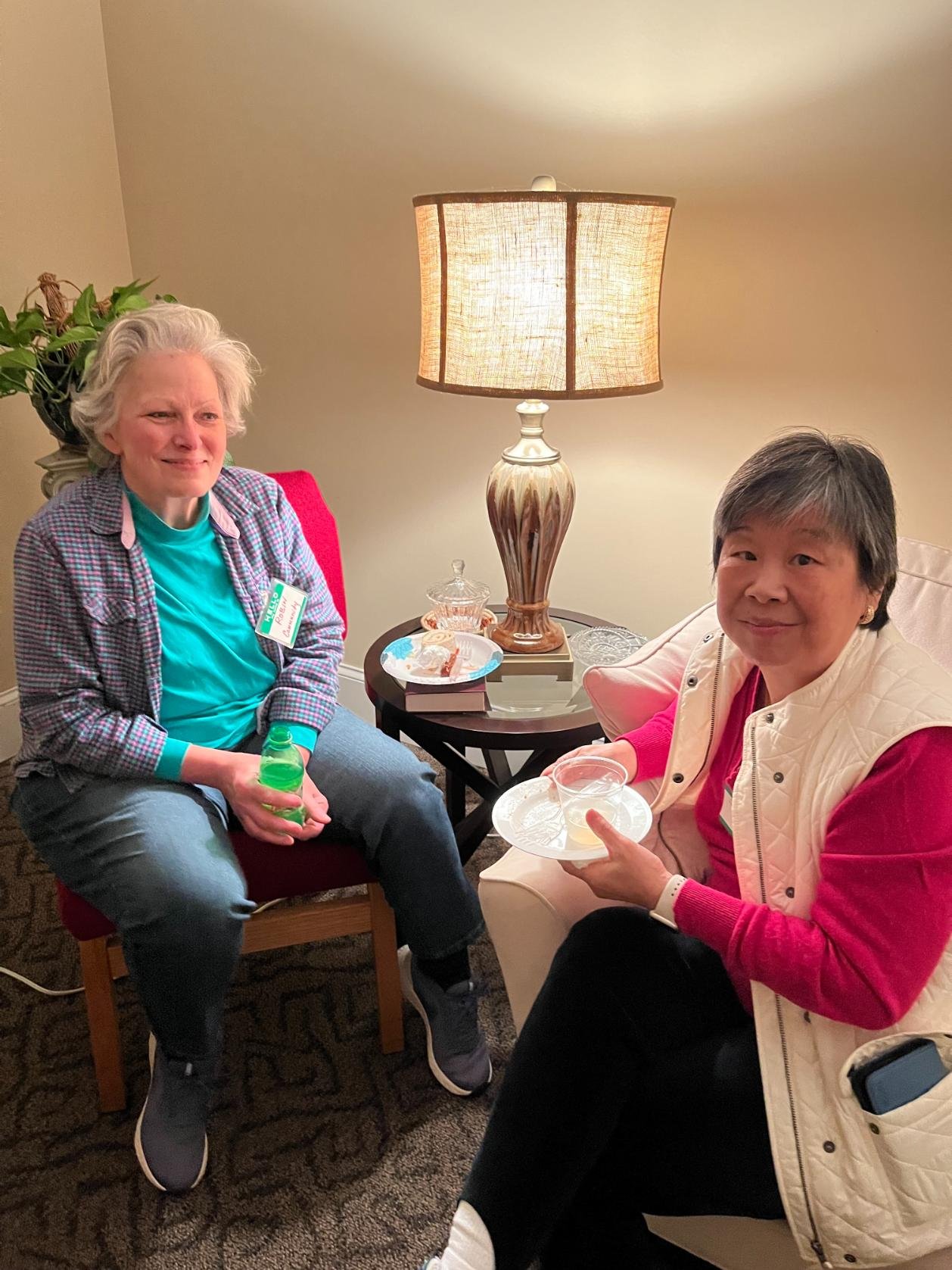 Two women sitting in a cozy room, one holding a glass of beverage and the other holding a plate with food, with a table and a lamp between them.