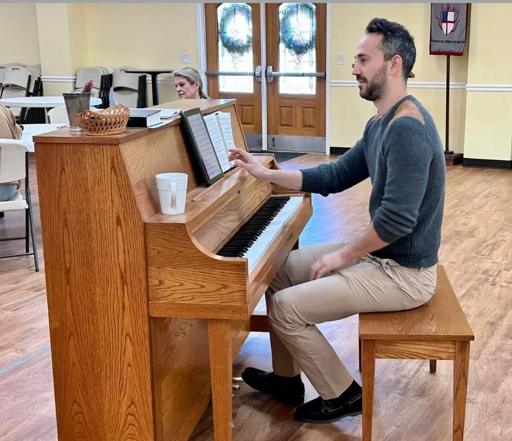 A man playing the piano in a room, with a woman sitting at a table in the background.