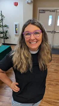 A woman with wavy hair and purple glasses smiling in an indoor setting.