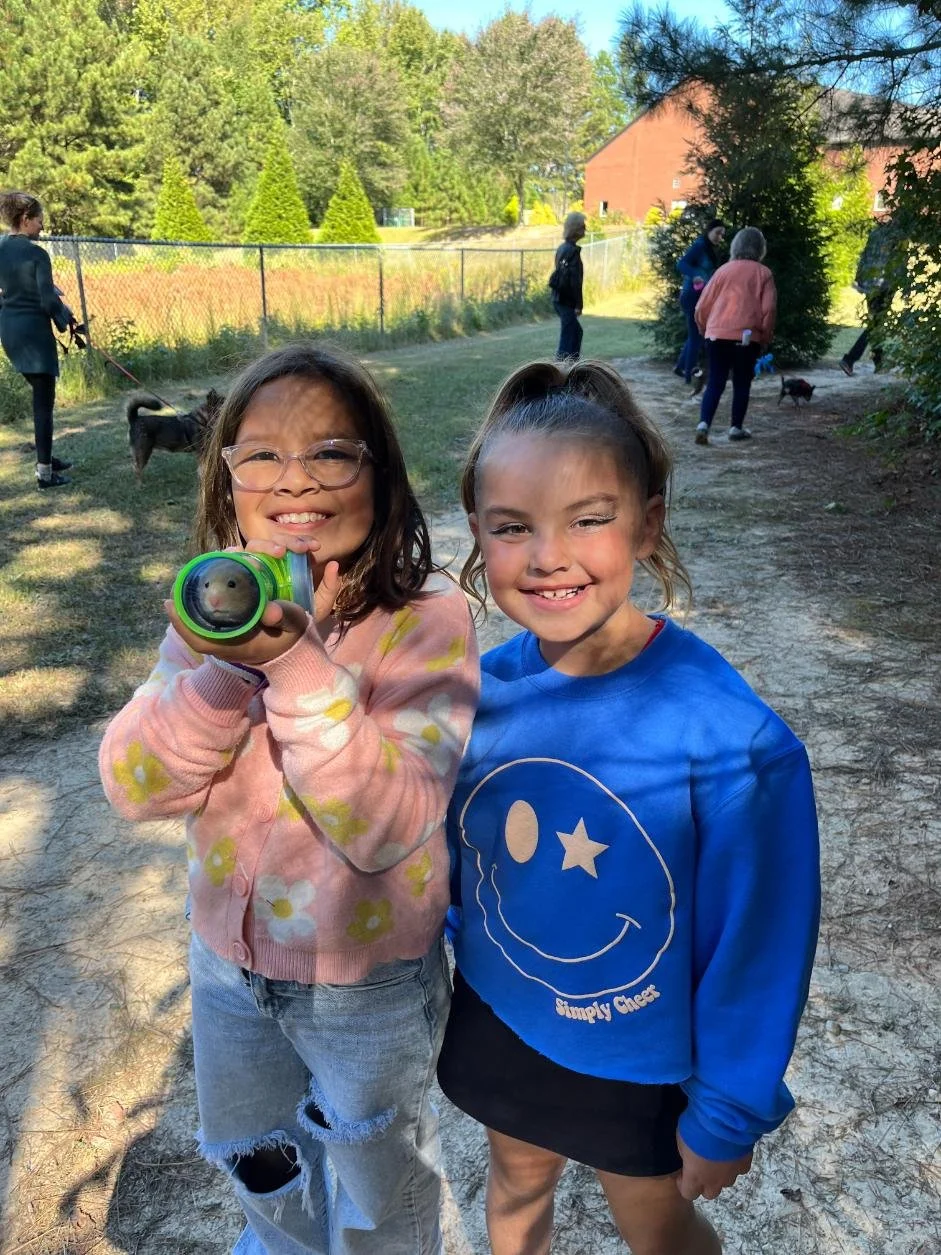 Two young girls smiling and posing for a photo outdoors in a park. The girl on the left is wearing glasses, a pink sweater with floral patterns, and ripped jeans, holding a toy binoculars. The girl on the right is wearing a blue sweatshirt with a smi
