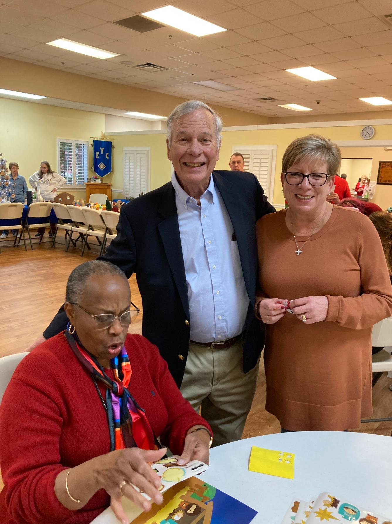 Three people at a social gathering, two standing and one seated at a table. The woman seated is looking at a Halloween-themed card with a surprised expression, while the man and woman standing behind her are smiling at the camera. The background show