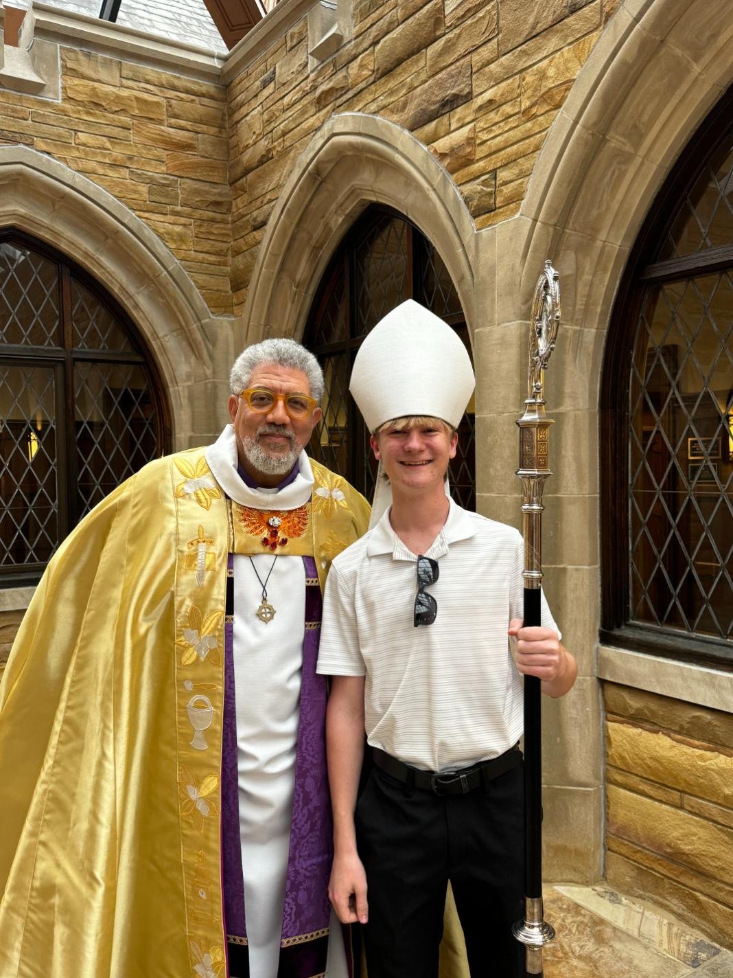 Two men standing in front of a stone building. One dressed in religious ceremonial robes with a yellow cloak and a cross necklace. The other in a white shirt, black pants, sunglasses, and a tall white hat, holding a staff. They are smiling.