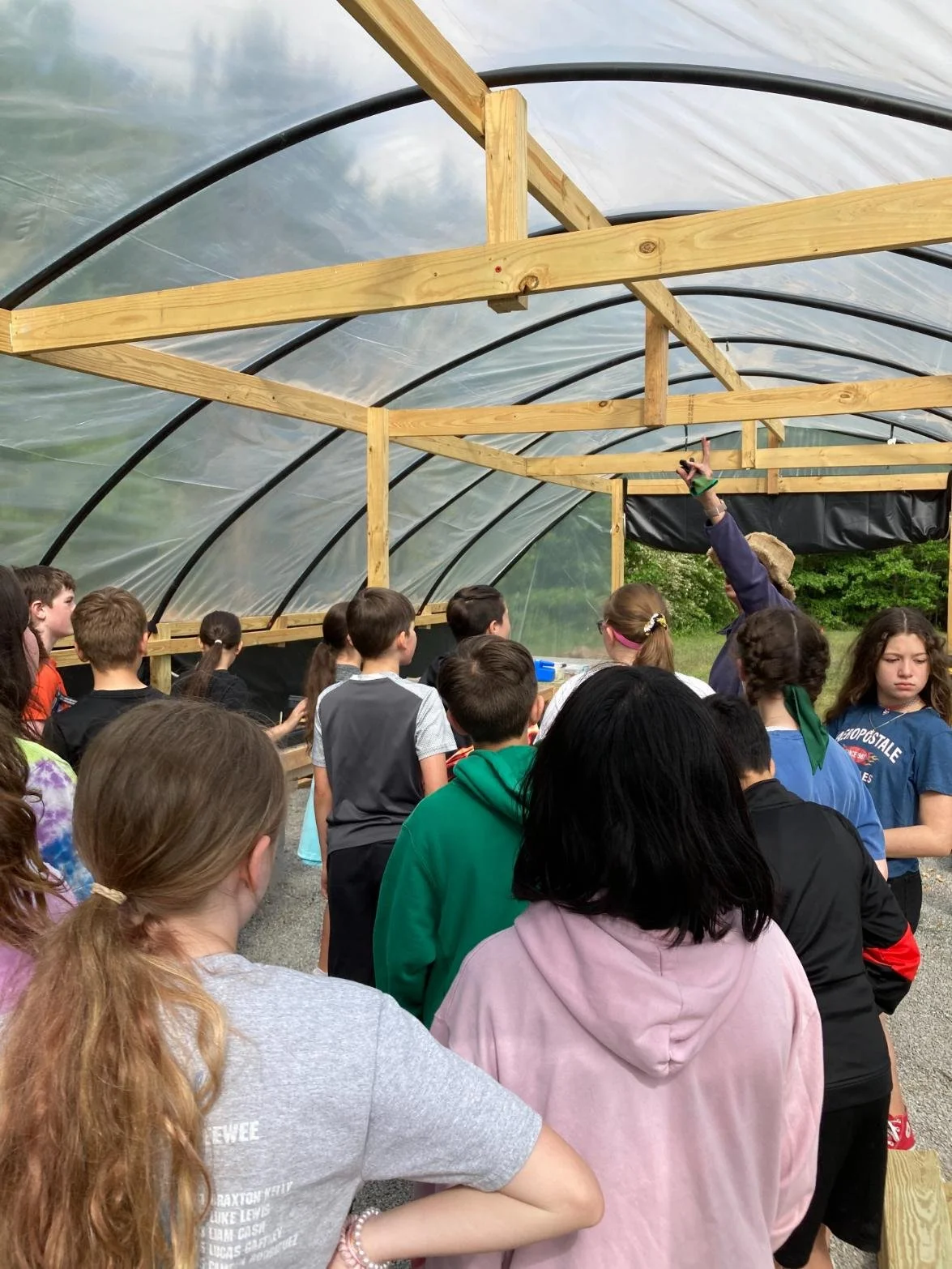 Group of children inside a greenhouse, listening to a woman giving a presentation.