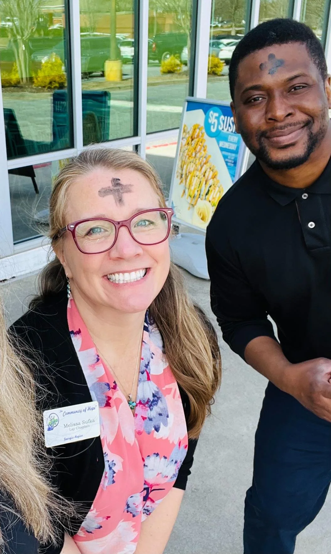 Smiling woman with glasses and a tattoo on her forehead, standing outside a building with glass doors, with a man beside her. There is a signboard for sushi and cars reflected in the glass behind them.