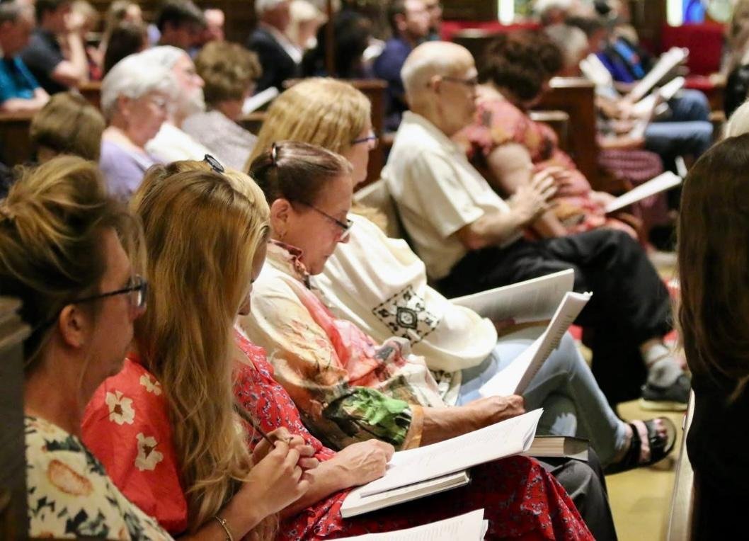 People seated in an audience, reading programs or booklets, attending an event or service, with women in the foreground wearing colorful blouses and glasses.