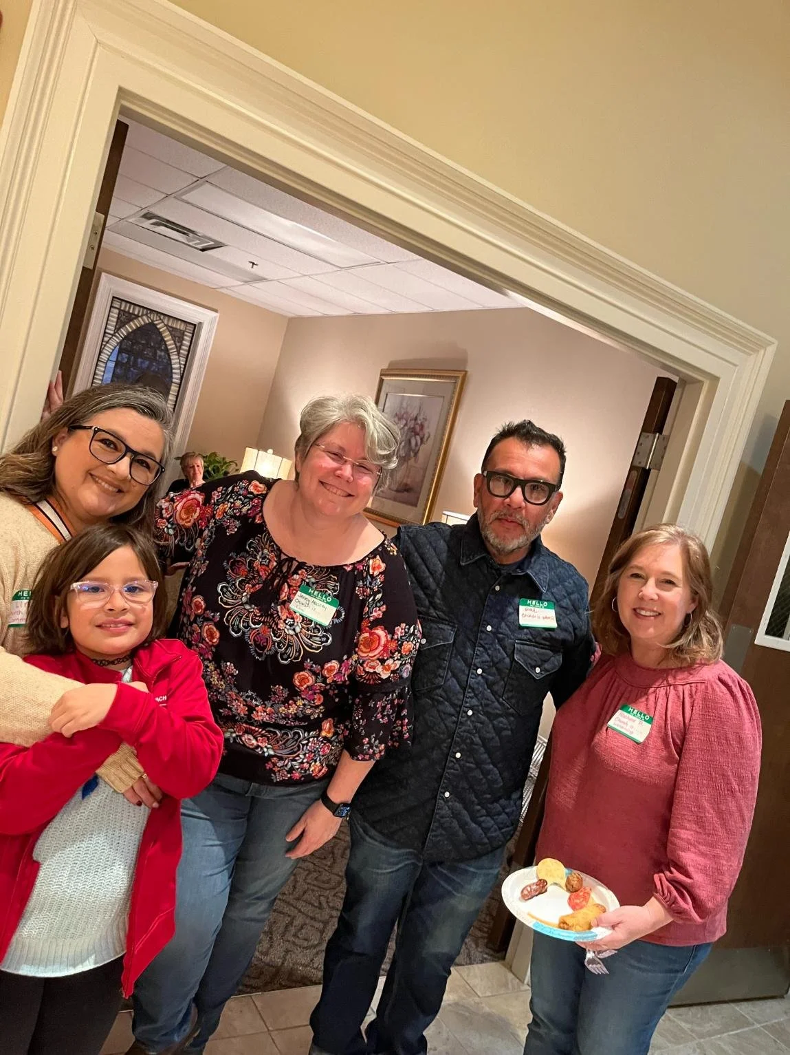 Group of five people at an indoor gathering, standing in a doorway, smiling for the camera. One woman is holding a plate with snacks.