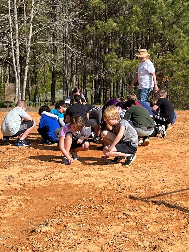 A group of children squatting and kneeling on a dirt clearing in a wooded area, participating in an outdoor activity with a guide. The background features dense trees and greenery.