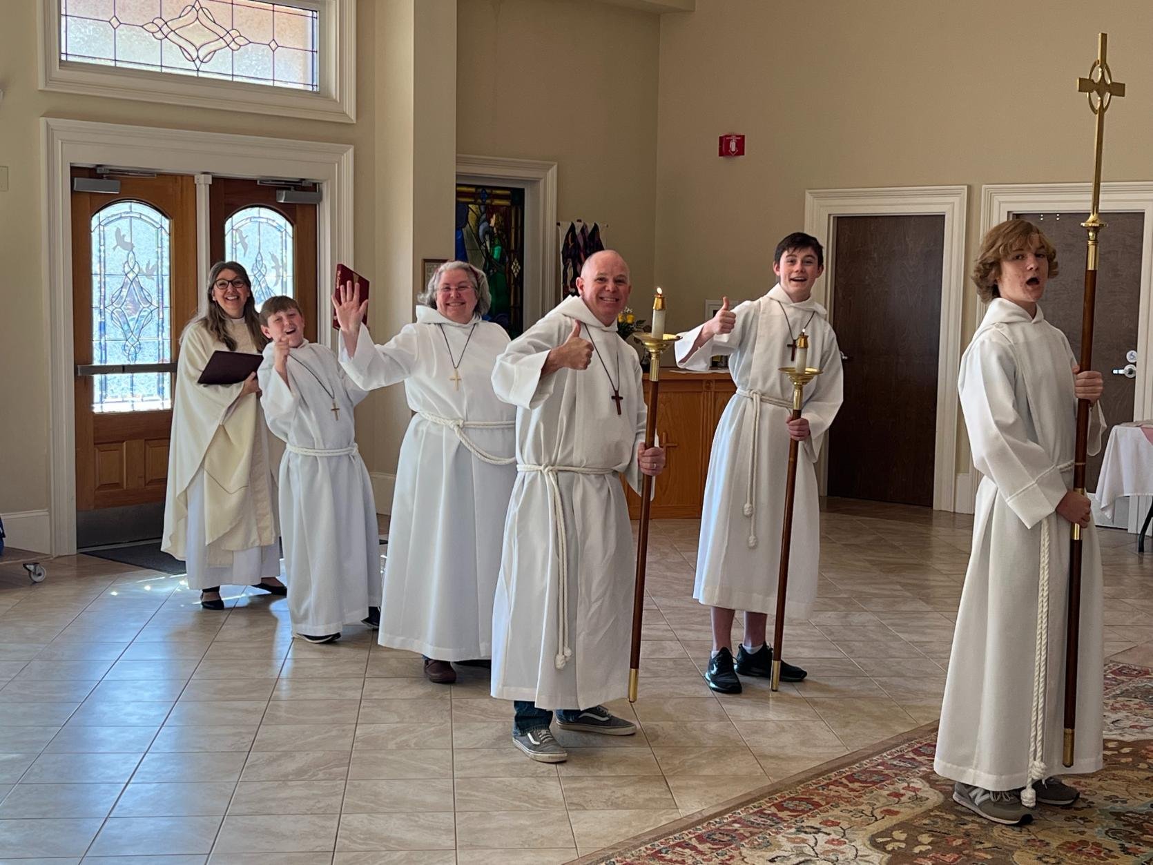 Group of people dressed in white religious robes, some holding candles and a staff, inside a church or chapel with stained glass windows and wooden doors, smiling and waving.