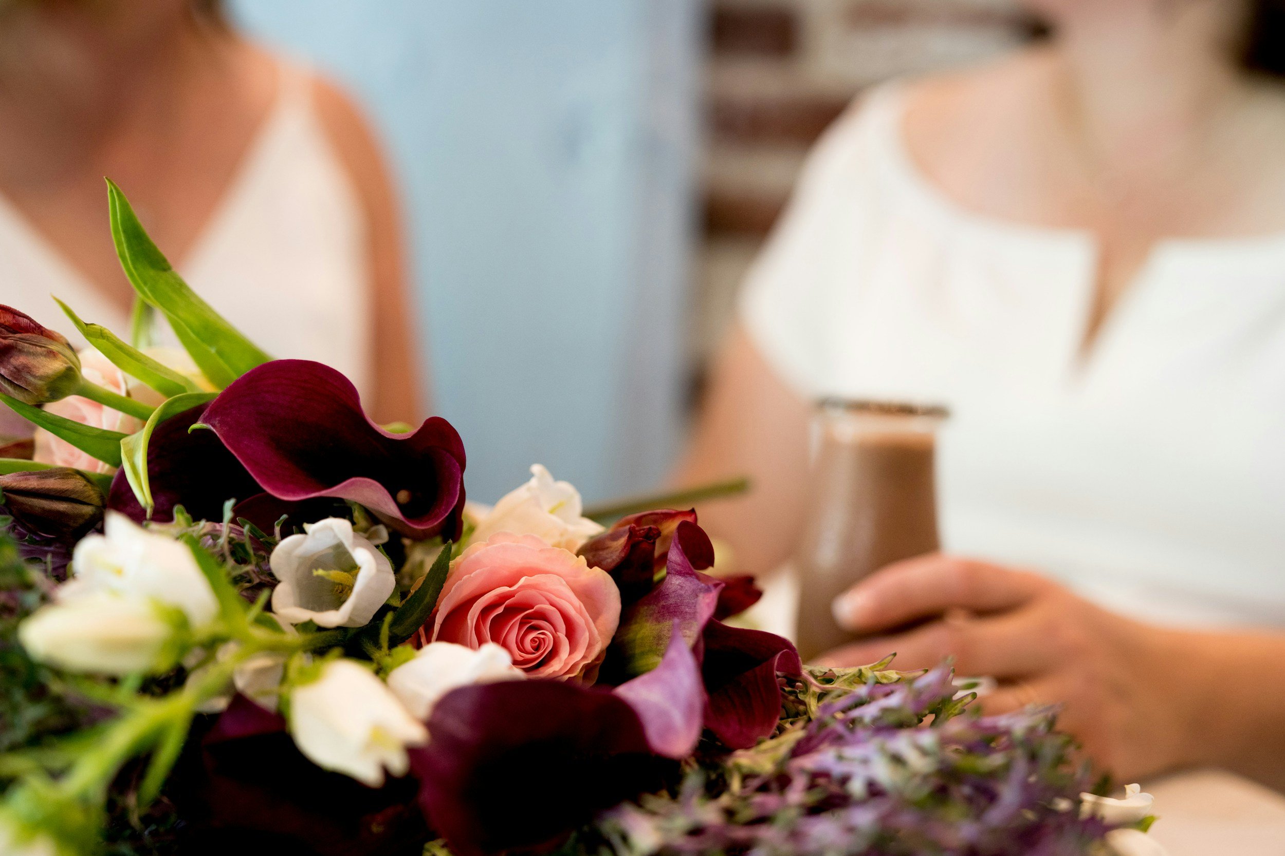 Close-up of a mixed bouquet of flowers including pink roses, dark purple calla lilies, white flowers, and greenery, on a wooden table in an indoor setting.