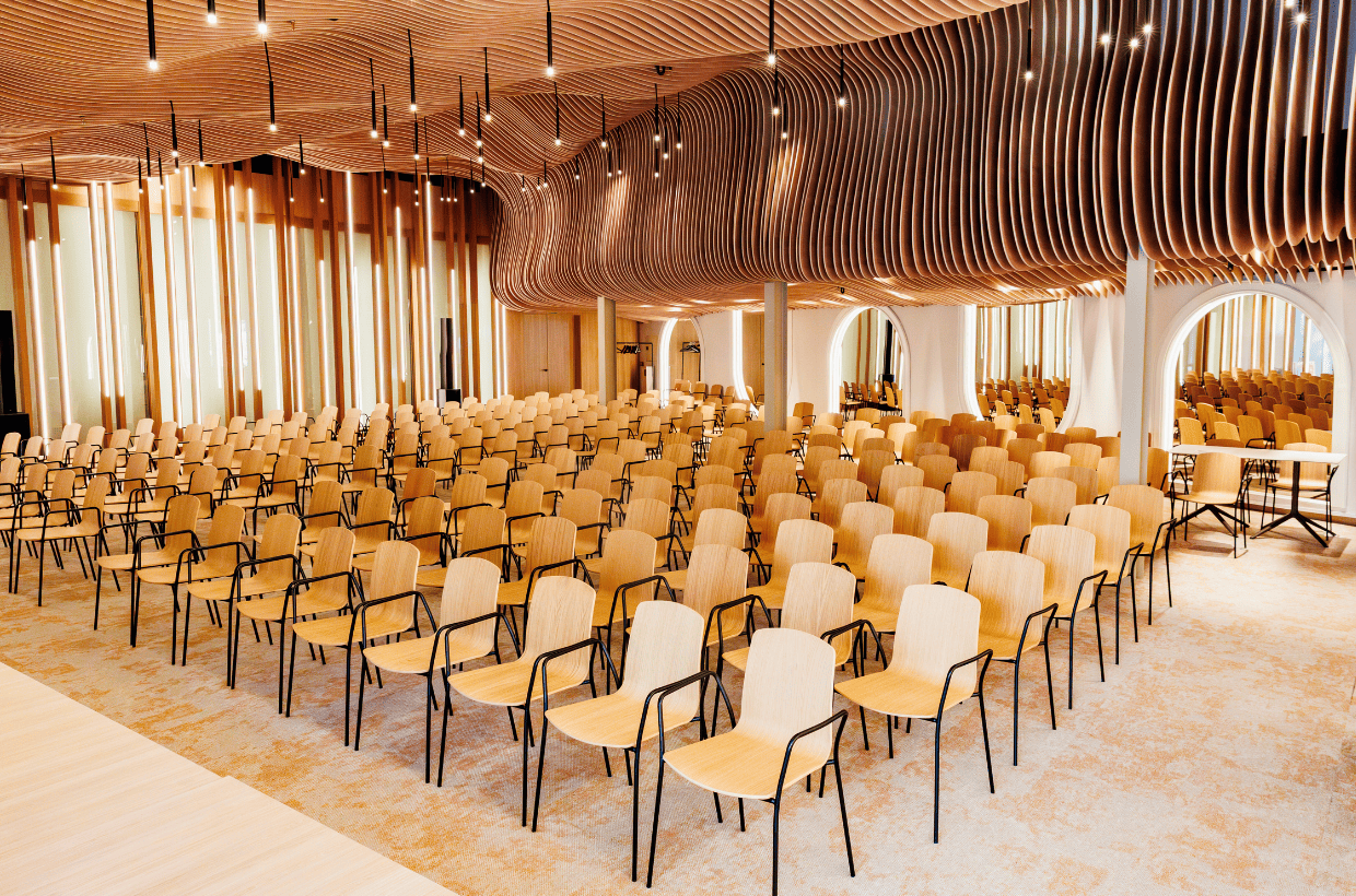 An auditorium with rows of wooden chairs with black armrests, a curved wooden ceiling with hanging light fixtures, and large arched windows along the side, allowing natural light to fill the space.