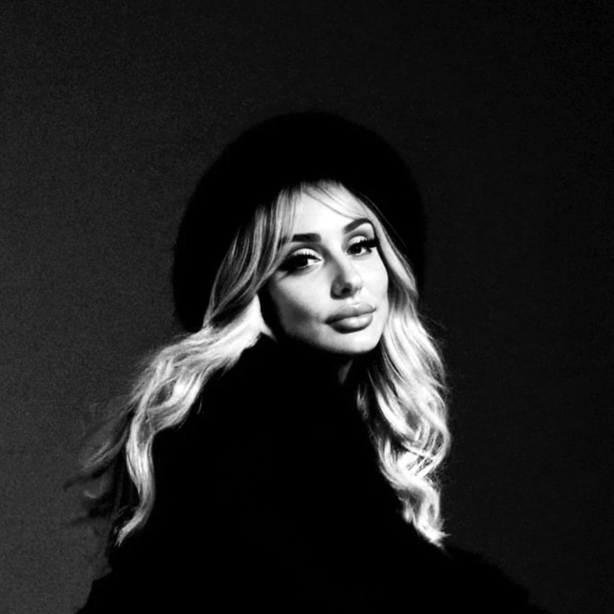 A black and white portrait of a woman with long wavy blonde hair, wearing a dark hat and dark clothing, looking at the camera