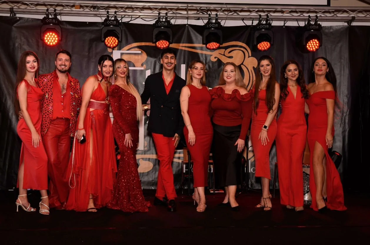 Group of nine people dressed in red and black, standing on stage with red lighting overhead, posing for a photo at an event or celebration.