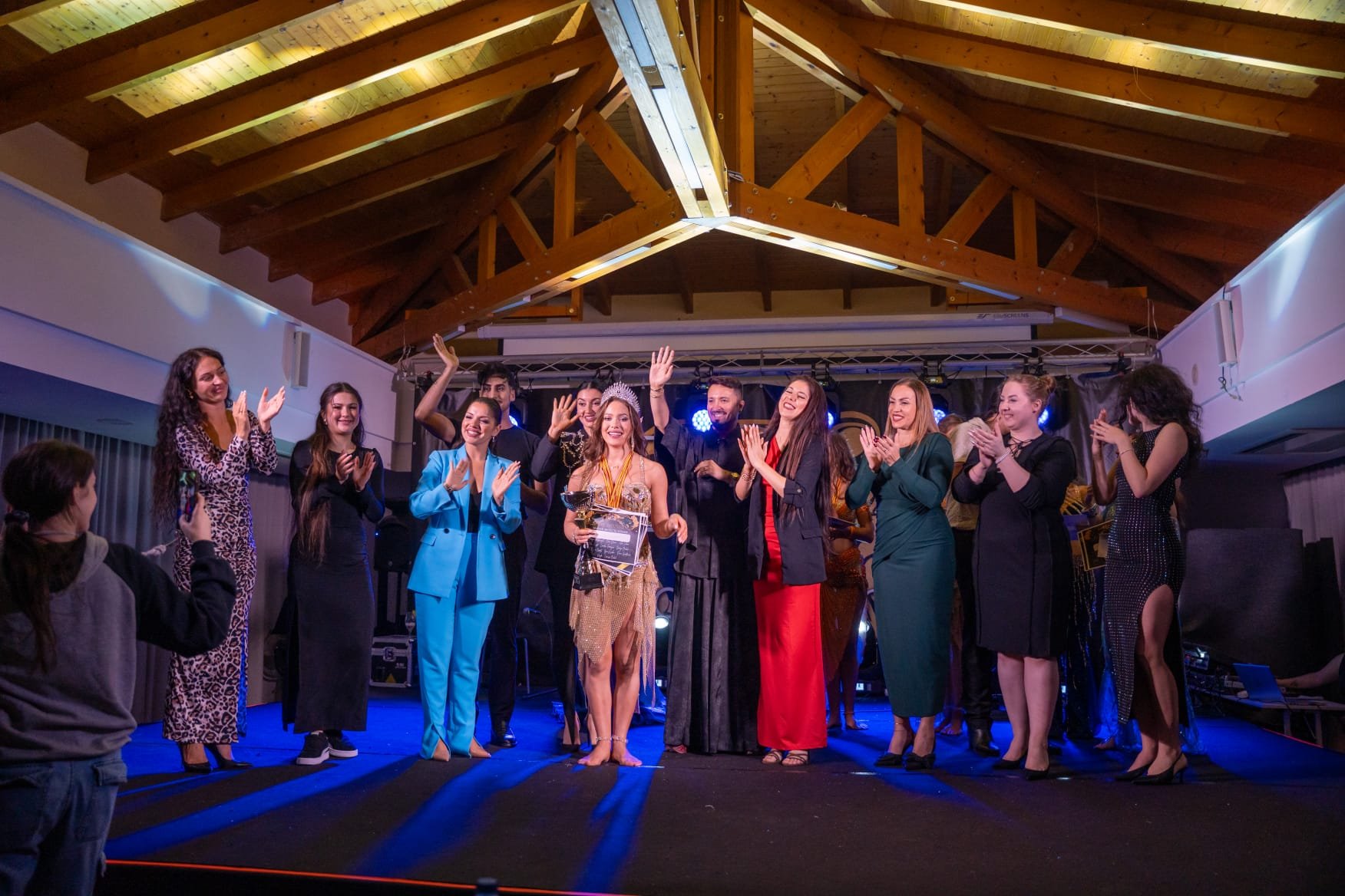 Group of people on stage celebrating, with some holding awards and trophies, applauding and smiling, in a venue with wooden ceiling beams and stage lighting.