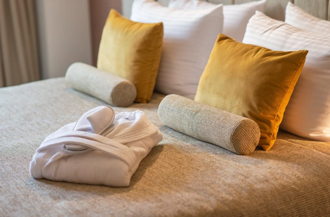 A neatly made bed with two white pillows, two mustard-colored pillows, two cylindrical bolsters, a folded bathrobe, and a towel in a hotel room.