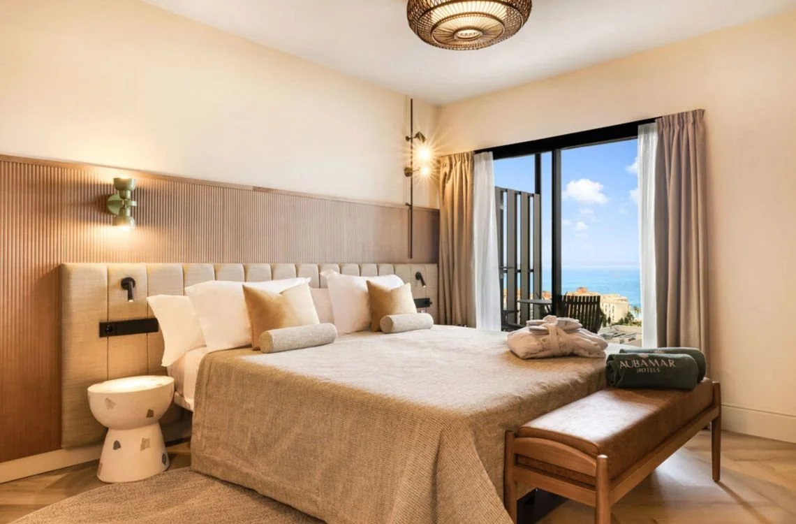 A beachside hotel room with a large bed, beige bedding, two cylindrical pillows, and a bench at the foot of the bed. There's a sliding glass door leading to a balcony with an ocean view, and the room has beige curtains, a wooden headboard, and a ceiling light.