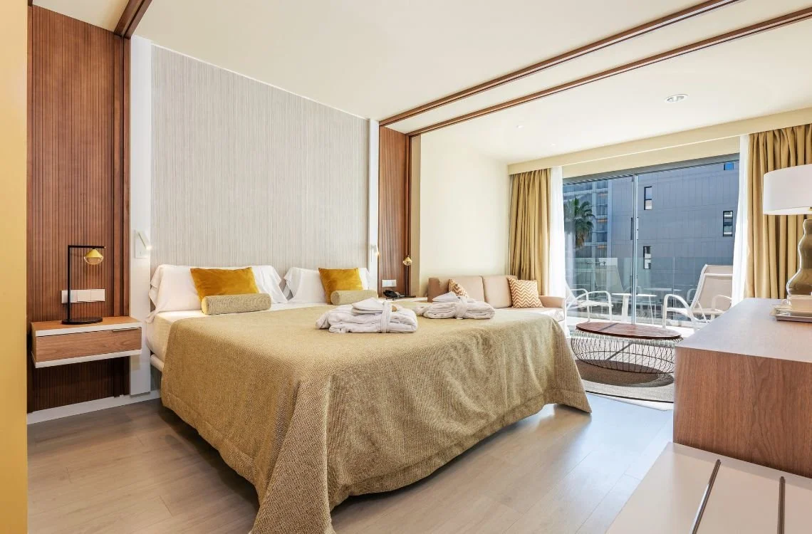 A modern hotel room with a king-sized bed, beige bedding, and four pillows, two yellow and two gray, with bathrobes on the bed. There is a bedside table with a lamp, seating area near large glass doors, and a balcony with outdoor furniture. The room has wood accents and light-colored walls.