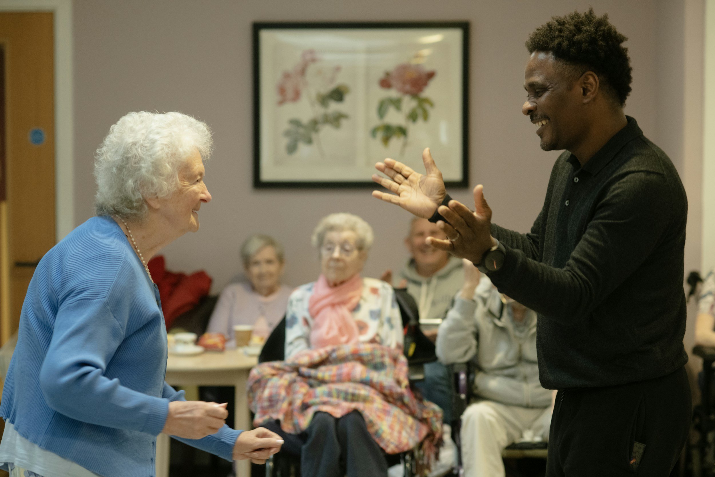 A young man and an elderly woman smiling and engaging in conversation in a social setting, with several elderly people watching and smiling in the background.
