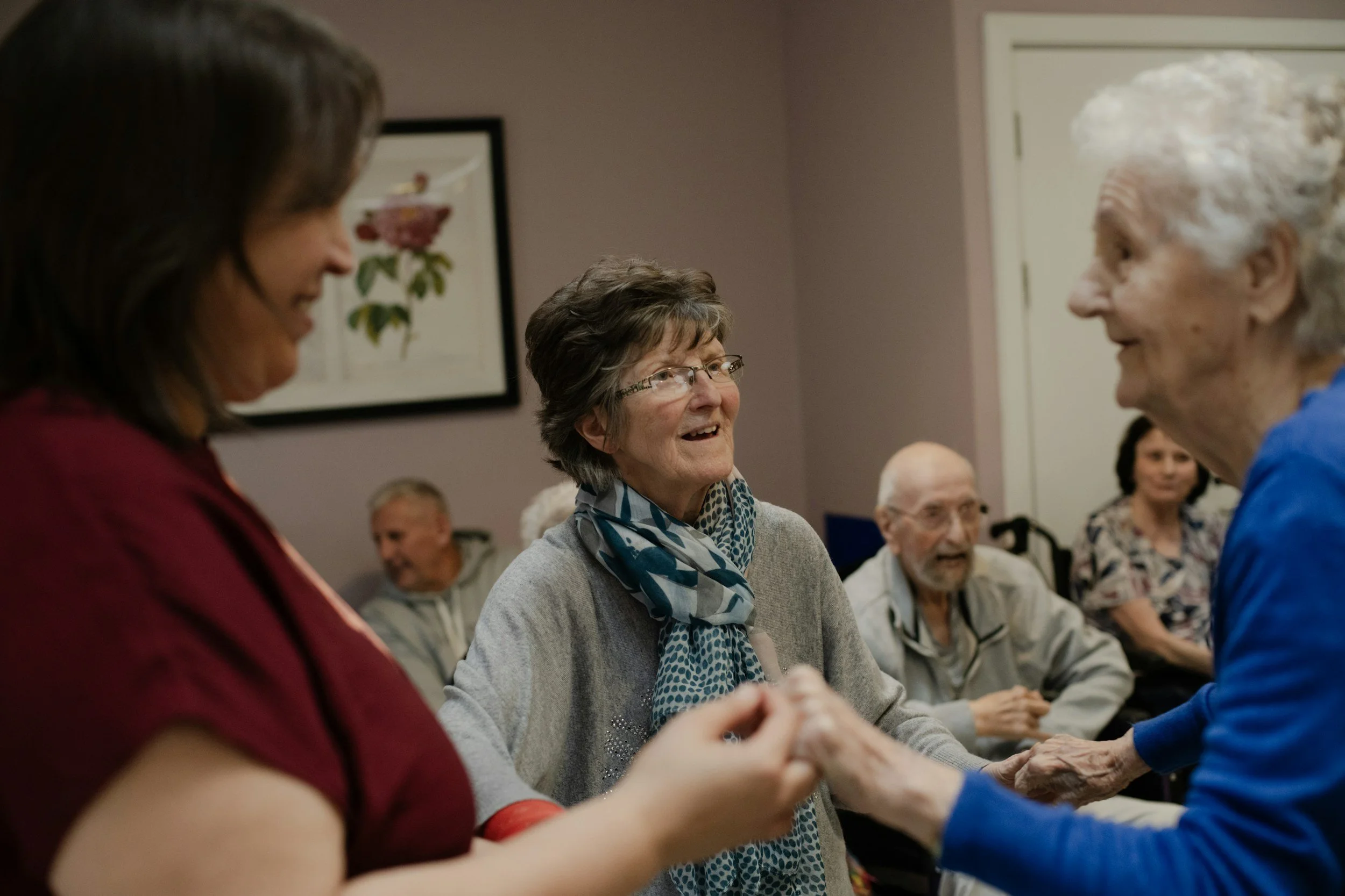 A group of elderly people and caregivers smiling and holding hands in a social gathering in a community room.