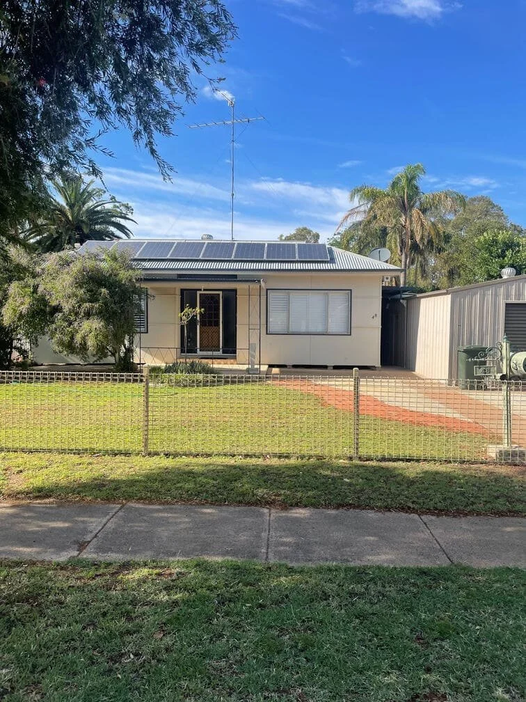 A small cream-colored house with solar panels on the roof, a front door with a screen, a large window with closed blinds, and a black side door. The house is surrounded by a grassy yard with a small garden, a tree on the left, and a metal fence. There are palm trees and other tall trees in the background, with a clear blue sky overhead.