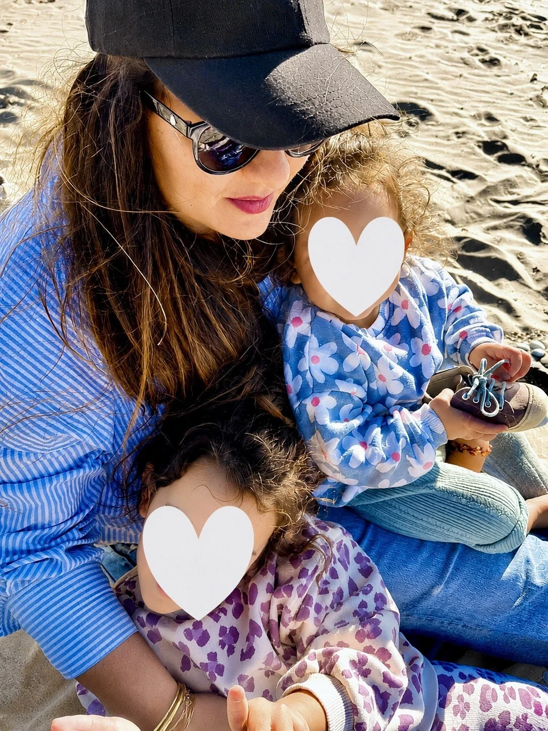Une femme et deux enfants assis sur la plage, tous portant des vêtements décontractés. La femme porte un chapeau noir, des lunettes de soleil et une chemise à rayures bleu. Elle tient un des enfants, qui porte un vêtement avec un motif de fleurs violettes. L'autre enfant, avec des cheveux bouclés blonds et portant un vêtement à motifs de fleurs blanches et roses, tient une chaussure. Les visages des enfants sont recouverts de cœurs blancs pour préserver leur vie privée.
