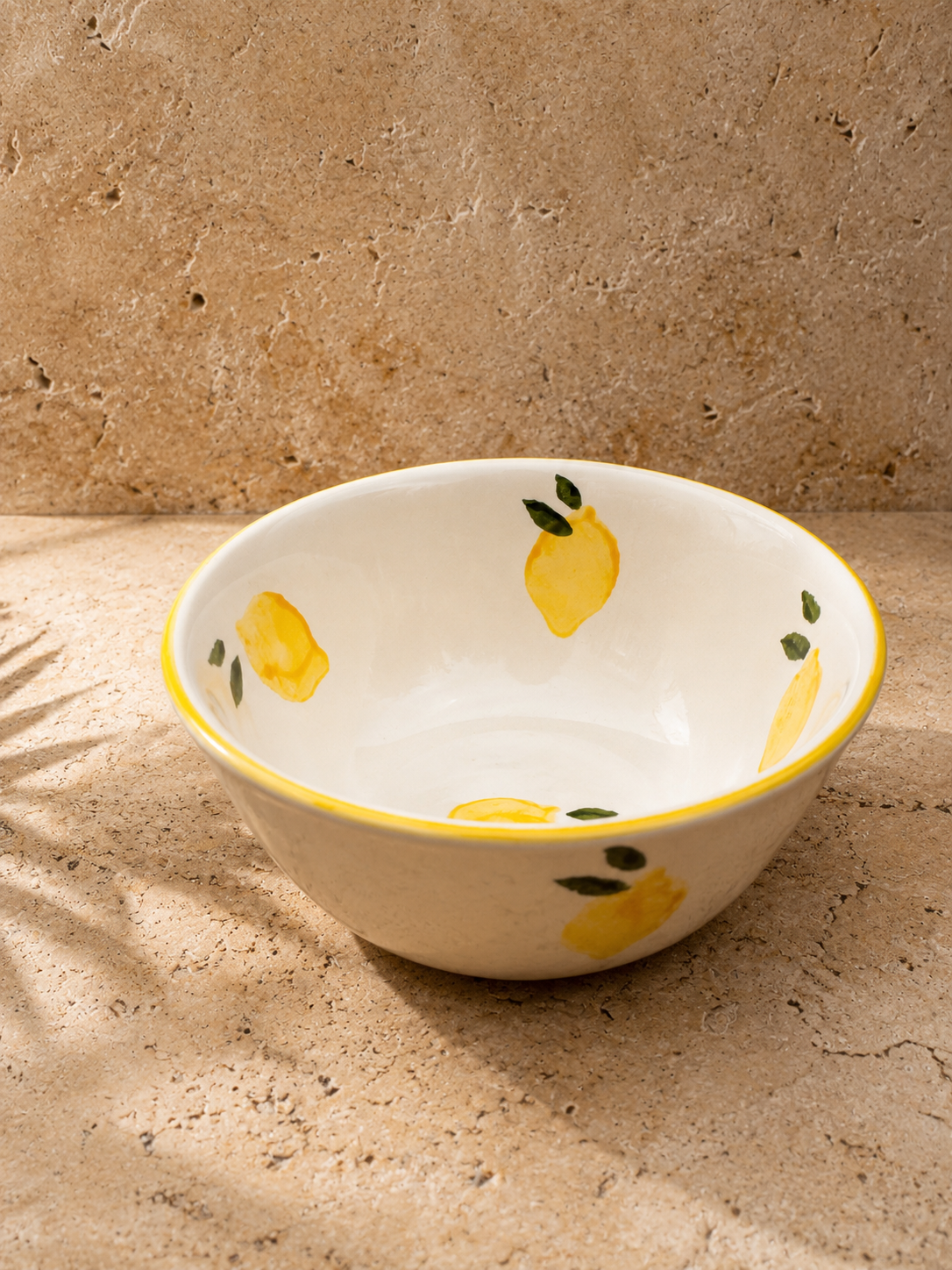 Bol en céramique blanc décoré de citrons jaunes et feuilles vertes, posé sur une surface en pierre ou en béton, avec un fond en pierre ou en béton et des ombres douces.