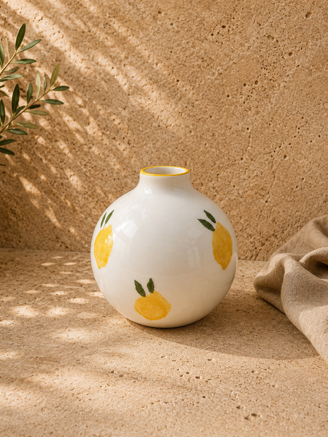 Vase en céramique blanche avec motifs de citrons jaunes et feuilles vertes, placé sur une surface en liège avec un fond de mur en liège et une branche d'olivier à gauche.
