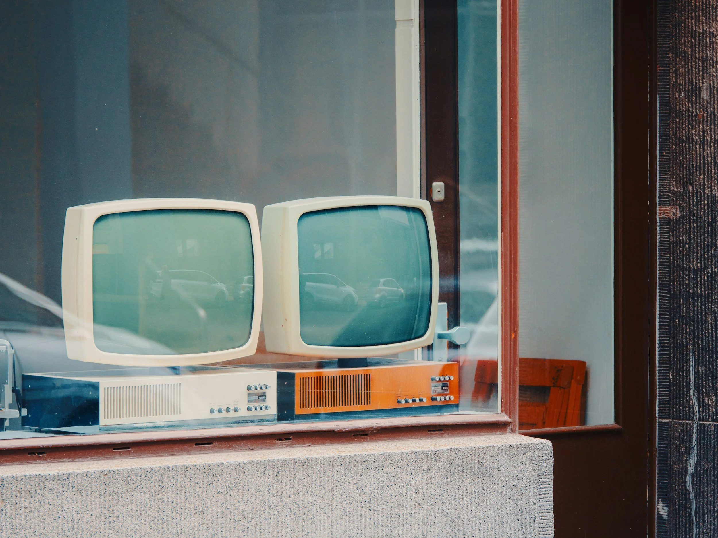 Two vintage computer monitors with curved screens on a display window ledge, reflecting parked cars outside.