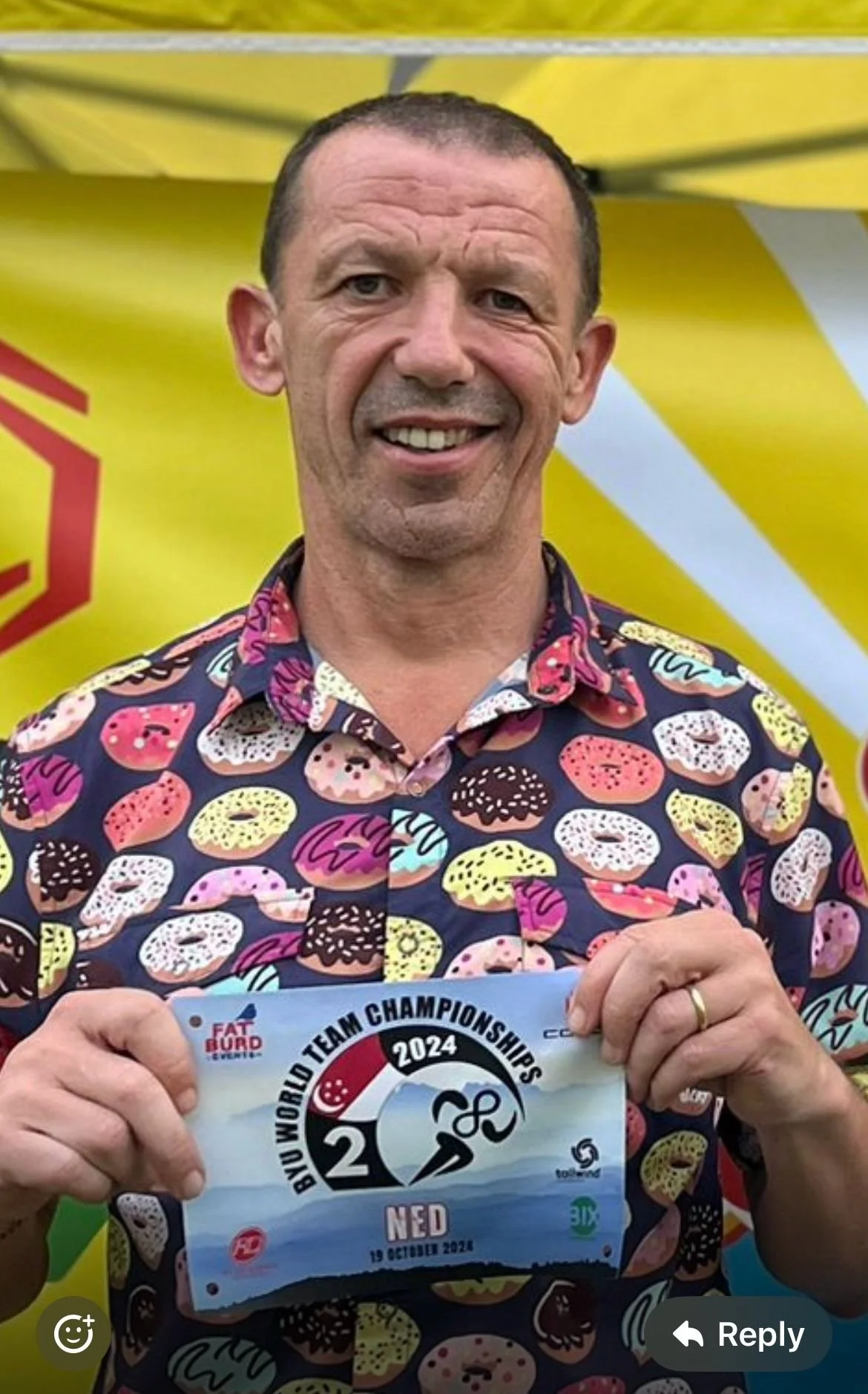 Man smiling and holding a ticket for the 2024 BIU World Team Championships, wearing a colorful donut-patterned shirt.