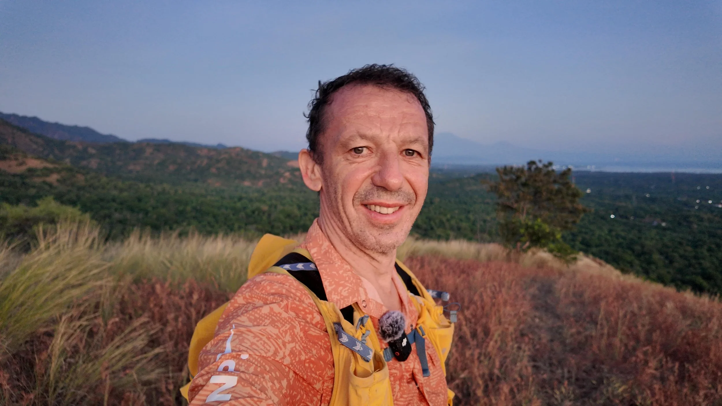 A man taking a selfie outdoors with a scenic landscape of hills, trees, and a body of water in the background during sunset.