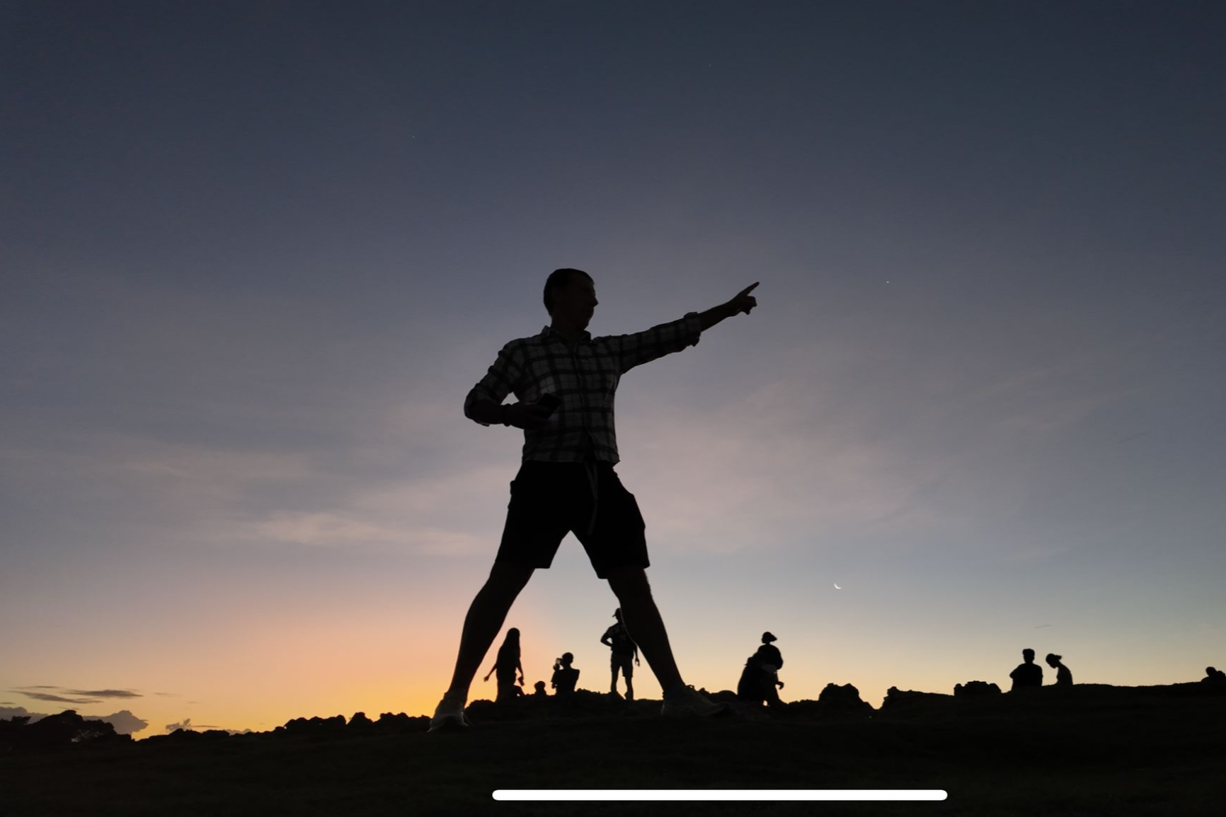 Silhouette of a man pointing with his right hand at sunset or dusk, with a group of people in the background on a rocky landscape.
