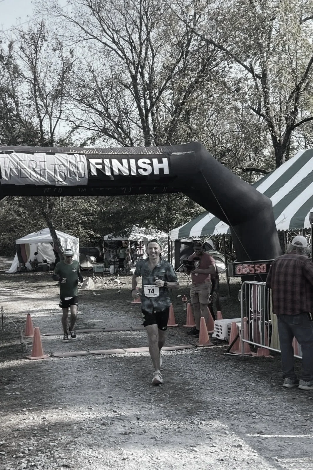 A man running through the finish line of a race, with two other runners behind him. The finish line is marked by a black archway with the word 'FINISH' on it. The setting appears to be an outdoor event with tents and trees in the background.