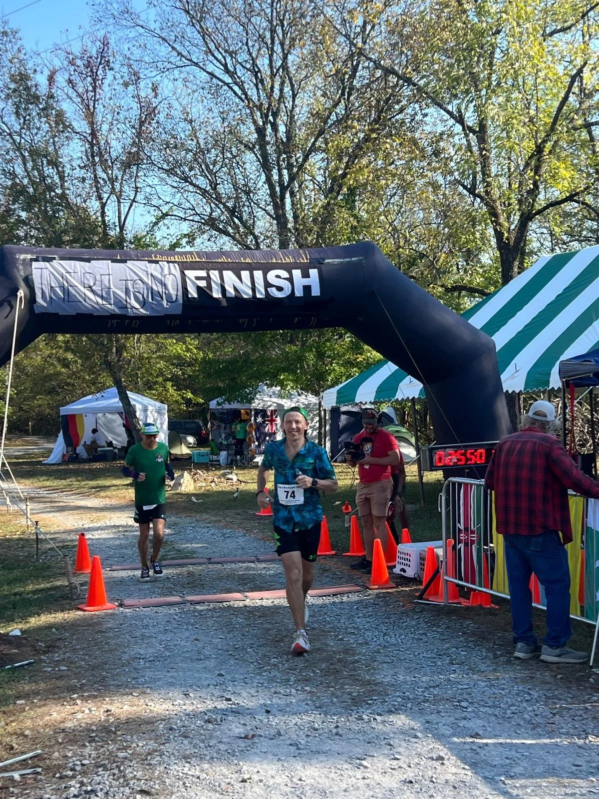 Runner finishes a race under a black finish line arch with a digital timer displaying 2:55:0, other participants, volunteers, and tents are visible in a park-like setting with trees.