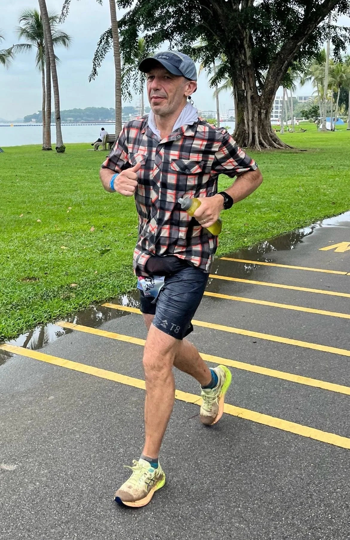 A man jogs on a wet path in a park near a body of water, wearing a checkered shirt, black shorts, running shoes, a cap, and a watch, holding a water bottle, with trees and a cloudy sky in the background.