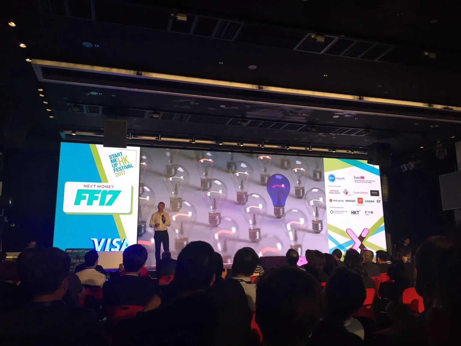 A speaker is standing on a stage at a conference, with a large screen behind him displaying lightbulb graphics and event branding. The audience sits in front of the stage in a darkened room.