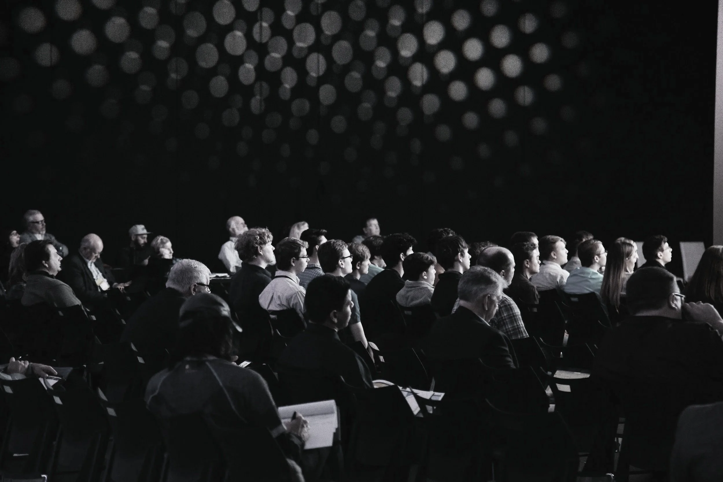 Audience attending a conference or seminar in a dark room, sitting in rows of chairs and listening to a speaker or presentation.