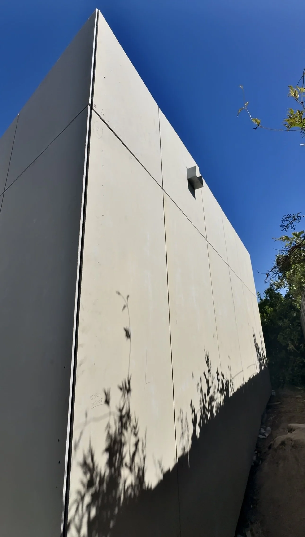 Modern building exterior with a sharp angled roof, white panels, and a small vent, casting shadows of nearby plants on the wall, against a clear blue sky.
