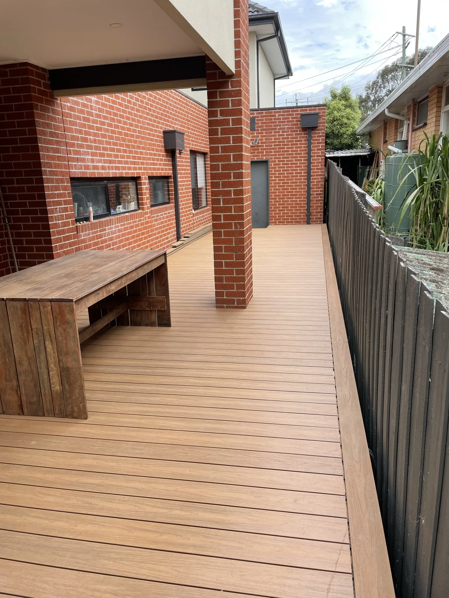 New wooden deck patio with a bench, enclosed by a dark fence, attached to a red brick house, with trees and a cloudy sky in the background.