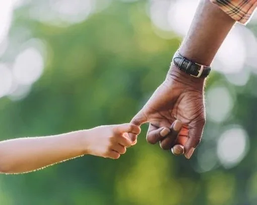 A child's hand holding an adult's hand outdoors with a blurred green background.