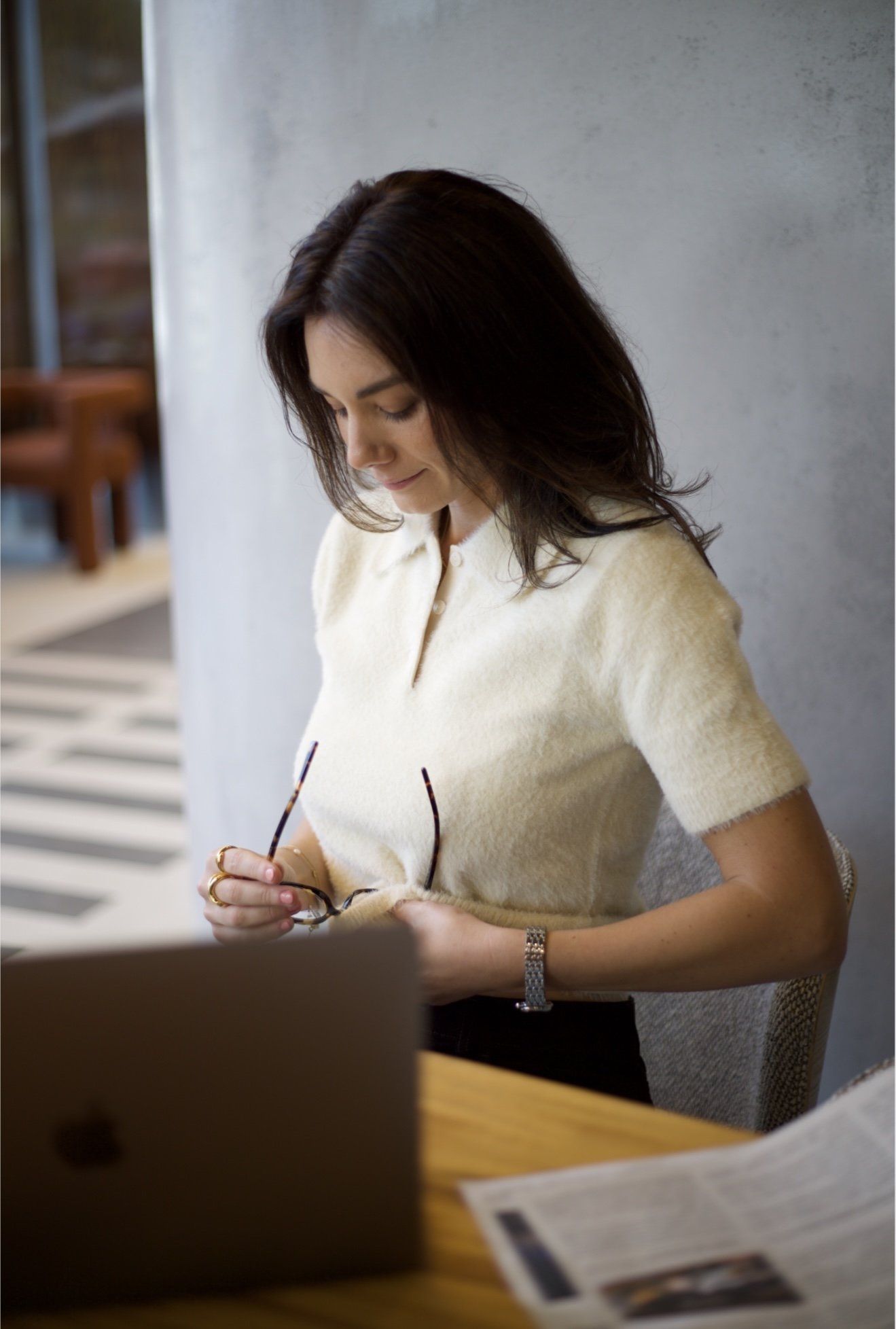 A woman with dark brown hair, wearing a cream-colored short-sleeve top, sitting at a table in a modern indoor space. She is holding glasses and appears to be looking down at something, with a laptop and a newspaper in front of her.