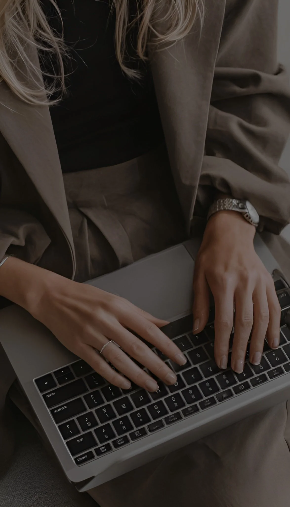Close-up of a woman typing on a silver MacBook keyboard, wearing a silver watch and rings, seated in a brown outfit.