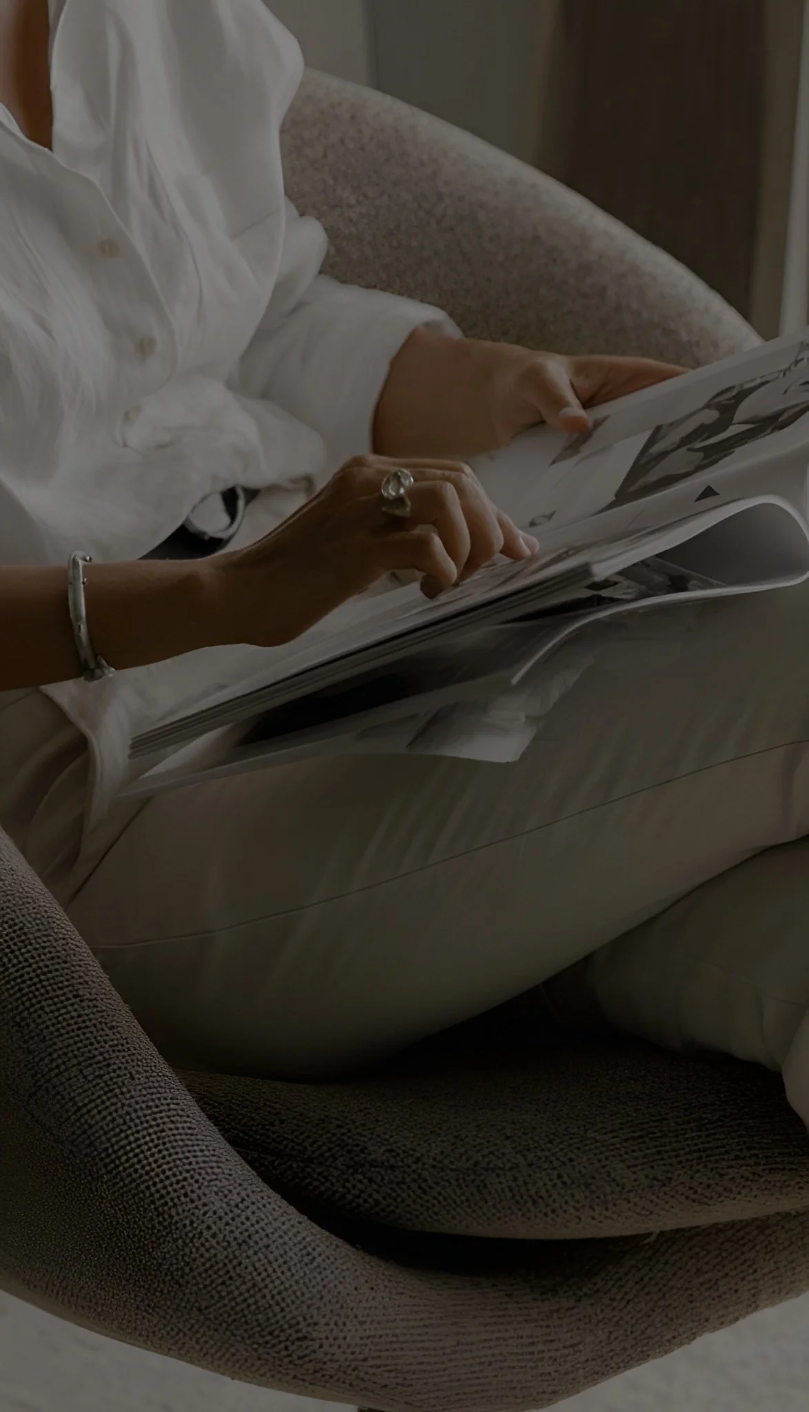 A person sitting on a beige chair, wearing a white shirt, holding and flipping through a magazine with black and white images.